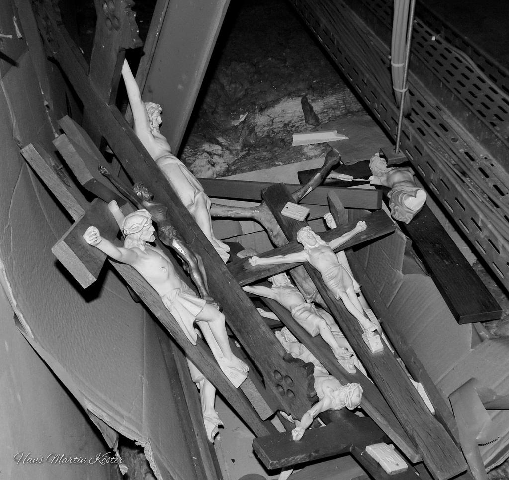 The black-and-white photograph shows several broken or discarded crucifixes lying haphazardly on the ground. Each crucifix features a figure of Jesus Christ nailed to the cross, with some figures partially detached or damaged. The crosses themselves are made of wood and appear to be scattered in a disordered pile. The scene evokes a sense of abandonment or destruction, possibly in a storage area or workshop. The lighting is dim, adding to the somber and reflective mood of the image.