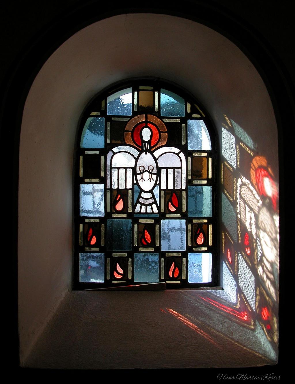 Das Bild zeigt ein farbenfrohes Kirchenfenster aus Buntglas, das in eine weiße, gewölbte Maueröffnung eingebettet ist. Das Fenster besteht aus vielen kleinen, rechteckigen Glasstücken in verschiedenen Farben, die ein symmetrisches Muster bilden.
Im Zentrum des Fensters ist eine stilisierte Taube dargestellt, die von einem runden Lichtschein umgeben ist. Unter der Taube befindet sich ein Kelch mit einer Hostie, ein klassisches christliches Symbol. Um diese zentrale Darstellung herum sind mehrere rote Tropfen- oder Flammensymbole angeordnet, die sich in den unteren Ecken des Fensters wiederholen.
Das Licht, das durch das Fenster fällt, wirft farbige Schatten auf die innere Fensterbank und den Boden, was dem Raum eine mystische und ruhige Atmosphäre verleiht. Die Szene wirkt meditativ und lädt zur Besinnung ein.