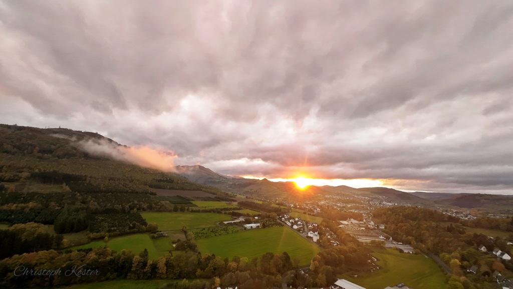 ENGLISH
This drone shot shows the view from Gierskopp towards Olsberg.
The sun is setting on the horizon. Lively clouds drift across the landscape. The Olsberg hut can be seen at the bottom left. A small cloud of fog hangs over Olsberg (left).
The picture was taken in October 2024.

DEUTSCH
Diese Drohnenaufnahme zeigt den Blick von der Gierskopp in Richtung Olsberg.
Am Horizont geht die Sonne unter. Lebhafte Wolken ziehen über die Landschaft. Links unten ist die Olsberger Hütte zu sehen. Am Olsberg (links) hängt eine kleine Nebelwolke.
Das Bild entstand im Oktober 2024