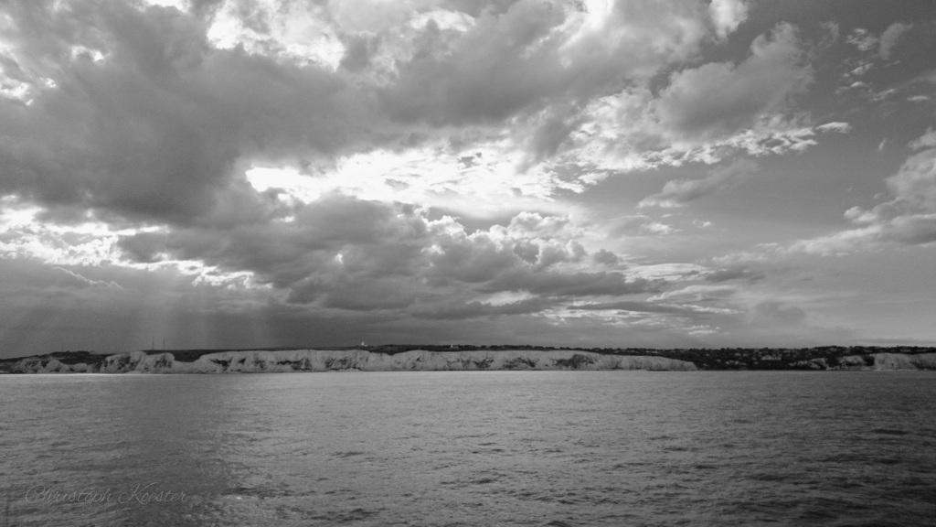 The black-and-white photograph captures a dramatic coastal scene. The foreground features a body of water, likely the sea, with gentle waves rippling across its surface. In the distance, a stretch of coastline is visible, characterized by prominent white cliffs that rise steeply from the water.
Above the cliffs, a sky filled with dense, dramatic clouds dominates the scene. The clouds vary in texture, with some areas appearing darker and heavier, suggesting the possibility of an impending storm. Streaks of light break through the clouds in places, adding contrast and depth to the image.
The overall mood of the photograph is moody and atmospheric, evoking a sense of the raw power and beauty of nature. The black-and-white tone enhances the dramatic effect, emphasizing the textures and contrasts within the scene.