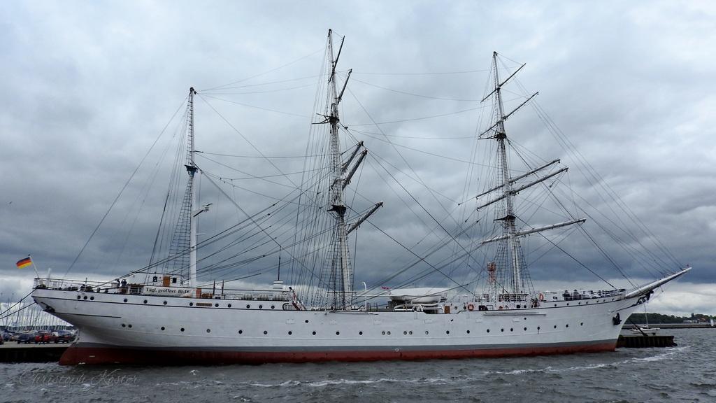 The image shows a large, three-masted sailing ship docked at a harbor. The ship has a white hull with a red waterline, and its tall masts are equipped with numerous ropes, rigging, and spars, typical of a traditional sailing vessel. The ship appears to be well-maintained and is flying a German flag at its stern.
The sky above is overcast with thick, gray clouds, suggesting a cloudy day. The water in the harbor is slightly choppy, with small waves visible. In the background, there are some industrial structures and cranes, indicating that the ship is docked in a port area. The overall scene conveys a sense of maritime history and the grandeur of classic sailing ships.