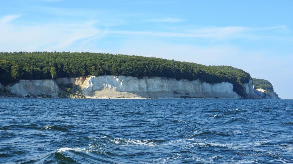The image showcases a stunning coastal landscape featuring towering white chalk cliffs. The cliffs rise dramatically from the sea, their bright white color contrasting sharply with the deep blue of the water below. The top of the cliffs is lined with dense, green forest, adding a touch of lush vegetation to the otherwise stark, rocky terrain.
The sea is slightly choppy, with waves creating texture on the water's surface. The sky above is clear and blue, with a few wispy clouds scattered across it, suggesting a pleasant, sunny day. The overall scene conveys a sense of natural grandeur and tranquility, highlighting the beauty of coastal geography. The image evokes a feeling of awe and admiration for the majestic cliffs and the vastness of the sea.