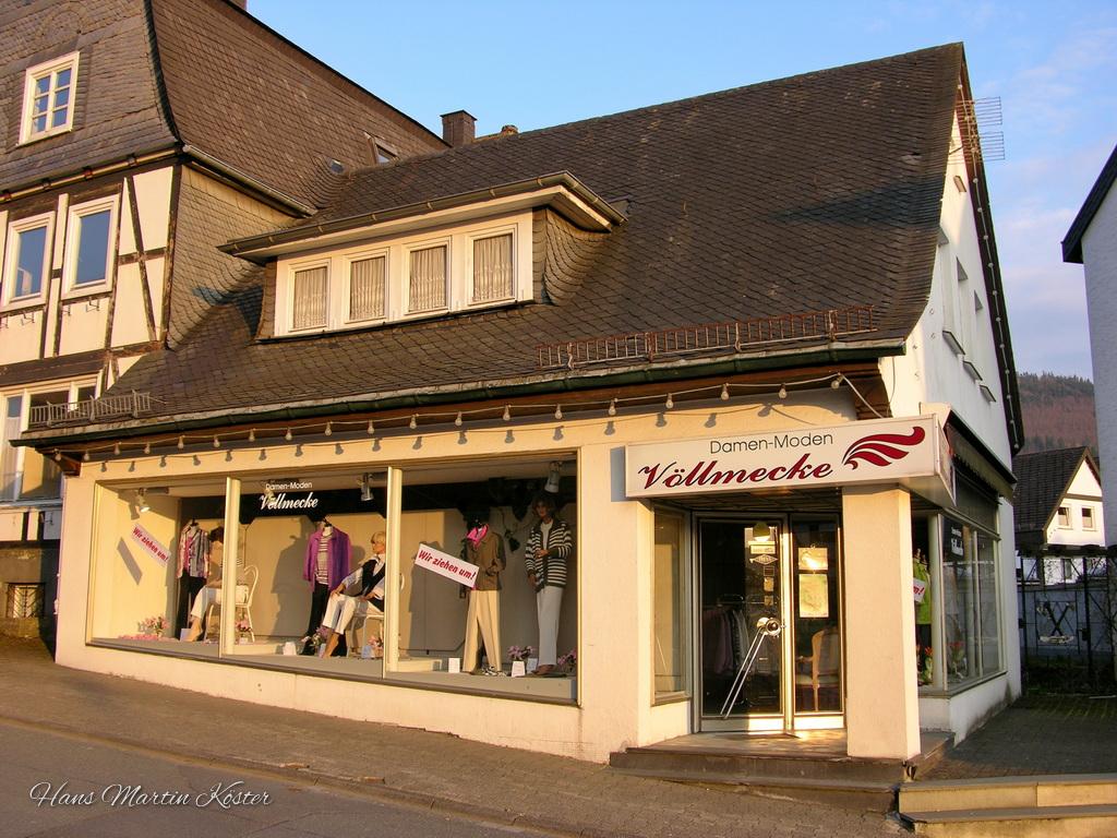 Das Bild zeigt die Fassade eines Ladengeschäfts in einer kleinen Stadt oder einem Dorf. Das Geschäft trägt den Namen "Damen-Moden Völlmecke", was auf den Verkauf von Damenmode hinweist. Der Laden befindet sich in einem Fachwerkhaus mit einem steilen, dunklen Schieferdach.
Die Schaufenster des Ladens sind groß und zeigen verschiedene Kleidungsstücke wie Jacken, Blusen, Hosen und Accessoires. Im Schaufenster sind auch einige Schilder zu sehen, darunter eines mit der Aufschrift "Wir ziehen um!" (Wir sind umgezogen!).
Die Fassade des Gebäudes ist hell gestrichen, und über dem Eingang hängt ein Schild mit dem Namen des Ladens und einem stilisierten Logo in Rot. Die Umgebung wirkt ruhig und typisch für eine kleinere Ortschaft. Der Himmel ist klar, und die Beleuchtung deutet auf eine frühe Abendstunde hin.