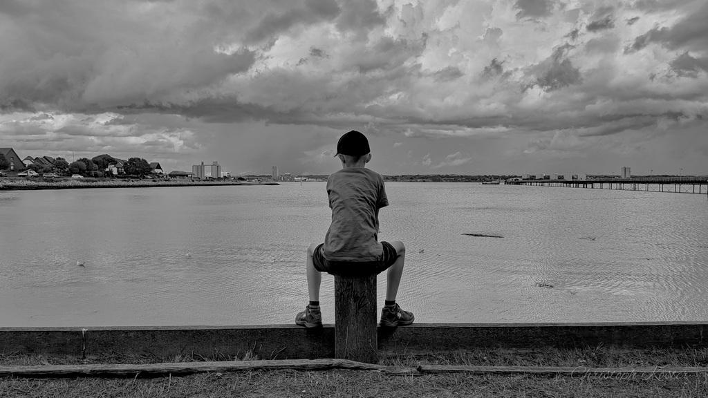 The thoughtful boy
Prospect Place Park, Hythe
July 2025

A black-and-white photograph showing a child sitting alone on a wooden post by a body of water. The child, wearing a cap, t-shirt, and shorts, is facing away from the camera, looking out over the water. The water is calm, and the horizon features a distant shoreline with buildings and trees. The sky is filled with dramatic, heavy clouds, suggesting an overcast or stormy atmosphere. The scene conveys a sense of contemplation and tranquility.