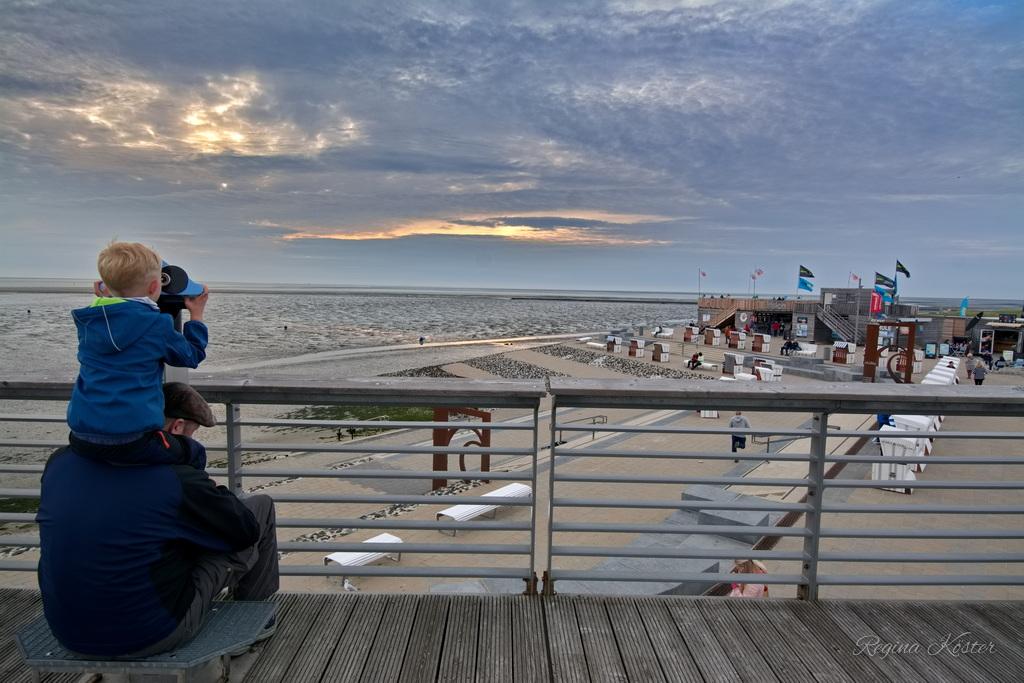 A color photograph capturing a serene seaside scene during what appears to be either sunrise or sunset. In the foreground, a man is sitting on a wooden bench on a boardwalk, holding a young child on his shoulders. The child is using binoculars to look out over the beach and the sea. The beach stretches out in the middle ground, with a few scattered beach chairs and small buildings or kiosks. Several flags are visible, fluttering in the breeze. The sea extends to the horizon, and the sky is filled with a mix of clouds, illuminated by the soft light of the setting or rising sun, creating a peaceful and contemplative atmosphere.