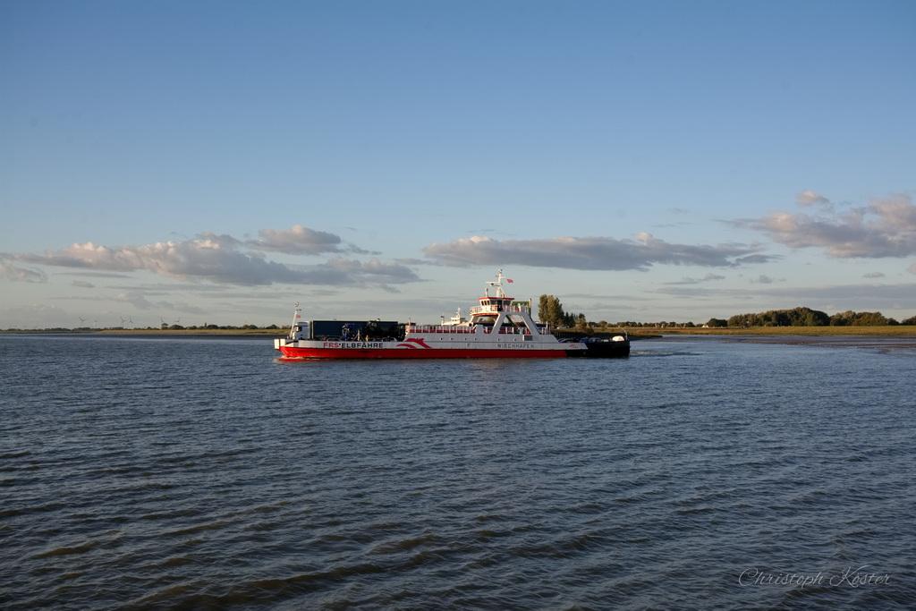 The image shows a red and white ferryboat sailing on calm waters. The ferry is centrally positioned in the frame, moving from left to right. The sky is mostly clear with some scattered clouds, and the sun is casting a warm light, suggesting either early morning or late afternoon. In the background, there is a flat, green landscape with a few trees and bushes along the horizon. The water is slightly rippled, reflecting the light from the sky. The overall atmosphere is peaceful and serene.