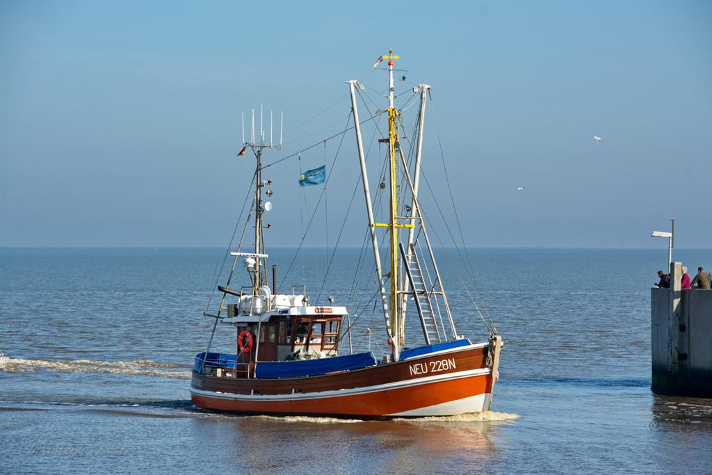 The image shows a fishing boat sailing in shallow waters, likely near a harbour or coastal area. The boat has a red and white hull with a blue cover over the rear deck. It is equipped with various fishing equipment, including antennas, ropes, and a small crane. The boat is moving from left to right, creating a gentle wake behind it.
In the background, the sea extends to the horizon under a clear blue sky. To the right of the boat, there is a concrete structure, possibly part of a pier or harbour wall, with a few people standing on it. The overall scene is calm and serene, with the boat appearing to be either leaving or entering the harbor.