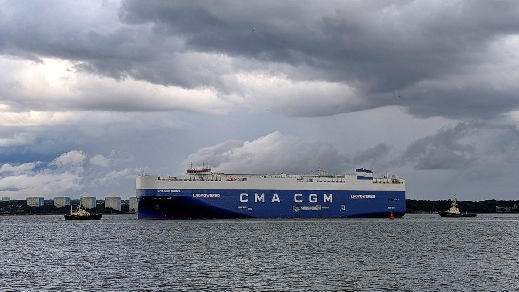 The image shows a large cargo ship named "CMA CGM Monza" sailing on a body of water. The ship is predominantly blue and white, with the letters "CMA CGM" prominently displayed in large white letters on the side of the hull. The words "LNG POWERED" are also visible, indicating that the ship is powered by liquefied natural gas.
The ship is accompanied by two smaller tugboats, one on each side, assisting it as it navigates through the water. The sky above is filled with dramatic, dark clouds, suggesting an overcast or stormy atmosphere. In the background, a city skyline with multiple buildings is visible along the horizon, indicating that the ship is near a port or coastal city. The water is relatively calm, with gentle waves.