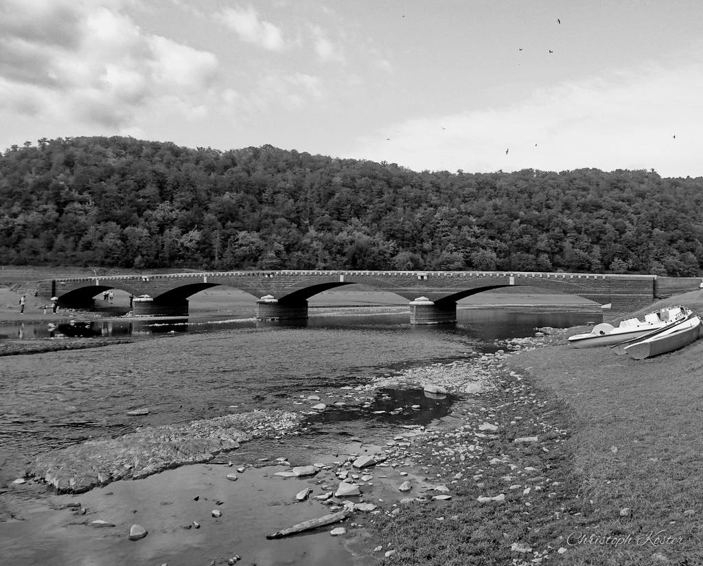 This black-and-white photograph depicts a serene riverside scene. In the foreground, there is a stony and partially dry riverbed with scattered rocks and shallow pools of water. Several small boats are beached on the shore.

Spanning the river is an old stone bridge with multiple arches, allowing water to flow underneath. The bridge appears sturdy and well-constructed, hinting at historical significance.

In the background, there are gently rolling hills covered with dense trees and vegetation. The sky above is partly cloudy, adding to the tranquil and timeless atmosphere of the scene.