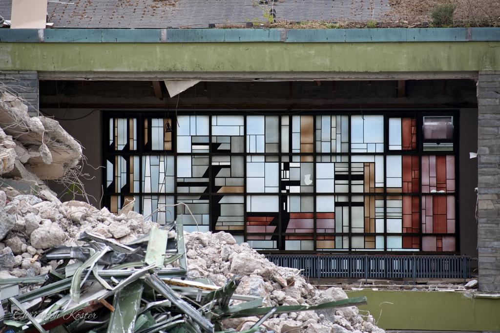 This image shows a partially destroyed building. In the foreground, there is a large pile of rubble consisting of broken bricks, concrete chunks, and twisted metal rods. The debris suggests significant structural damage.

Behind the rubble, you can see the remains of a building's facade. The structure has large, multi-paned windows, many of which are shattered or missing. The interior appears to be gutted, with visible damage to the floors and walls inside. The upper part of the building still has some intact sections, including a green-painted edge along the rooftop, but it looks unstable and heavily affected by whatever caused the destruction.

The scene evokes a sense of abandonment and damage, possibly from a natural disaster or demolition.