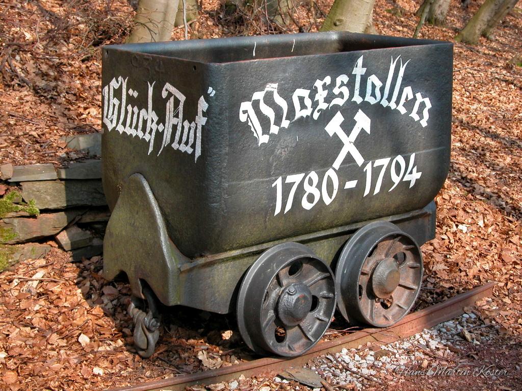 Das Foto zeigt einen alten, schwarzen Förderwagen aus dem Bergbau, der auf einem kurzen Stück Schiene im Wald steht. Der Wagen ist aus Metall und hat vier kleine Räder. Auf der Seite ist in weißer Schrift ‚Glück Auf‘ sowie ‚Maxstollen 1780–1794‘ zu lesen, daneben sind Schlägel und Eisen als Symbol abgebildet. Um den Wagen herum liegt braunes Laub, und im Hintergrund stehen mehrere Bäume und ein kleiner Steinhaufen. Die Szene wirkt wie ein historisches Bergbau-Denkmal oder Ausstellungsstück in der Natur.
