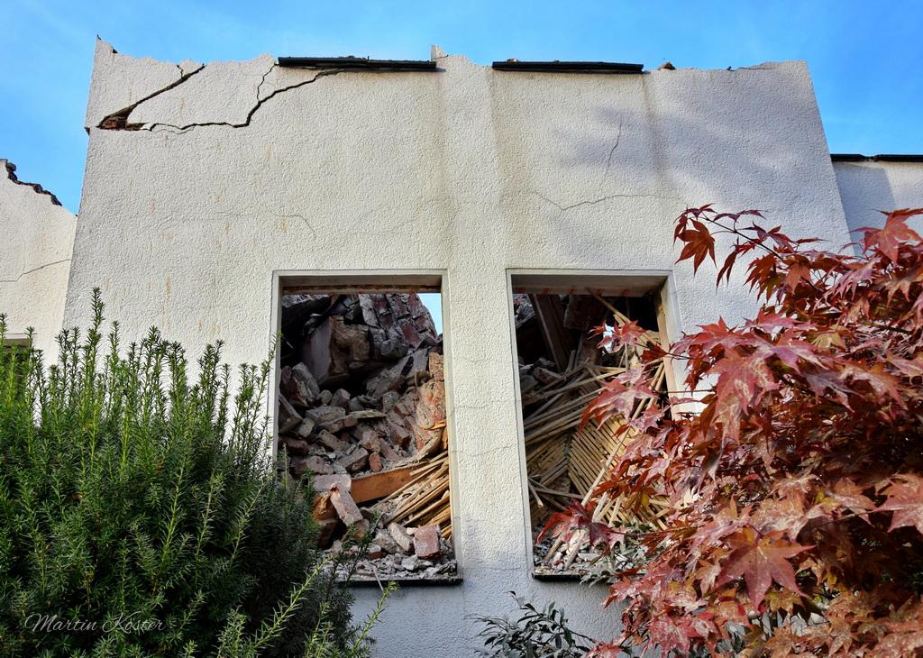 The image depicts a partially collapsed building, showing clear signs of severe structural damage. The focus is on two large, empty window openings in a weathered, off-white stucco wall. The windows are missing their frames and glass, revealing piles of rubble and debris inside—broken bricks, concrete chunks, and twisted metal rods, likely remnants of the building’s internal structure.

The wall itself appears aged, with visible cracks running vertically and horizontally, and patches where the stucco has crumbled away. The roofline above the windows is uneven, with sections of the overhang missing or damaged. In the foreground, green shrubs and plants grow along the base of the wall, adding a touch of nature to the scene. To the right, a branch with vibrant red autumn leaves partially frames the image, contrasting with the otherwise somber and dilapidated setting. The sky above is clear and blue, suggesting the photo was taken during the day.