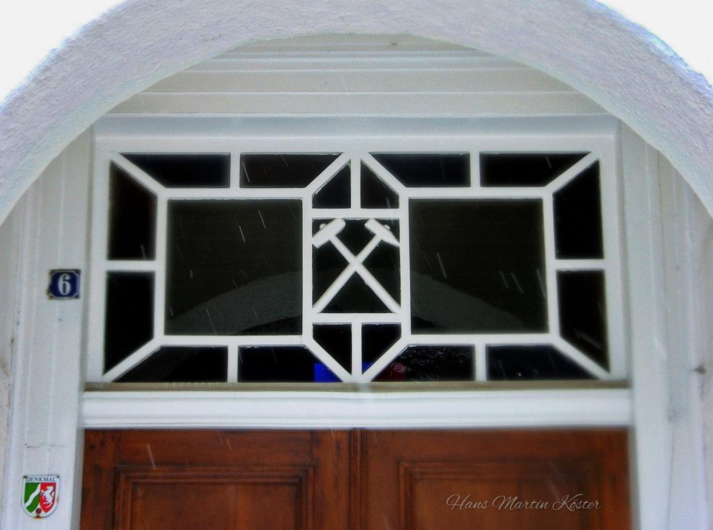 Das Bild zeigt den oberen Teil einer Eingangstür in einer weißen Hausfassade. Über der Tür befindet sich ein rechteckiges Fenster mit dekorativem, weißem Gitterrahmen, der in geometrische Formen unterteilt ist.

Im Zentrum des Fensters ist ein Symbol mit zwei gekreuzten Hämmern zu sehen, das an ein traditionelles Bergbauzeichen erinnert. Die Glasflächen sind teilweise dunkel getönt, sodass der Innenraum kaum erkennbar ist.

Links neben der Tür ist eine blaue Hausnummerntafel mit der Zahl 6 angebracht. Darunter befindet sich ein kleines Wappen mit grün-roten Elementen.

Der obere Bereich der Tür ist von einem weiß verputzten Rundbogen eingefasst. Am unteren Bildrand ist der obere Teil einer massiven Holztür mit warmer brauner Farbe sichtbar.

Feine helle Streifen im Bild deuten auf fallenden Regen oder Schnee hin. Die Szene wirkt ruhig und detailreich, mit einem Fokus auf architektonische Gestaltung und symbolische Elemente.
