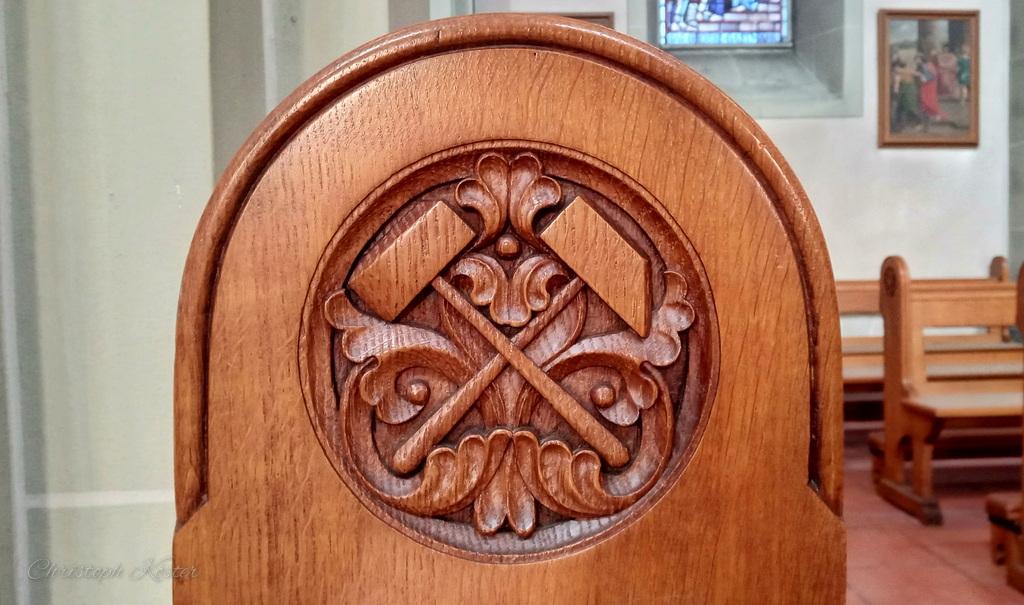 Das Bild zeigt eine Nahaufnahme der Rückenlehne einer hölzernen Kirchenbank. Das helle, glatt polierte Holz ist kunstvoll verziert und weist eine warme, honigbraune Farbe auf.

Im Zentrum der Lehne befindet sich ein kreisförmiges Schnitzrelief mit einem dekorativen Muster. In der Mitte sind zwei gekreuzte Werkzeuge dargestellt, die wie Hämmer oder Bergbaugeräte aussehen. Um das Symbol herum sind ornamentale Verzierungen mit geschwungenen Formen und pflanzlichen Motiven eingearbeitet.

Im unscharfen Hintergrund erkennt man den Innenraum einer Kirche oder Kapelle, mit weiteren Holzbänken, hellen Wänden und einem kleinen Fenster mit buntem Glas. An der Wand hängt zudem ein religiöses Bild.

Die Szene wirkt ruhig und detailreich und hebt die handwerkliche Kunst der Holzschnitzerei sowie die sakrale Atmosphäre des Raumes hervor.