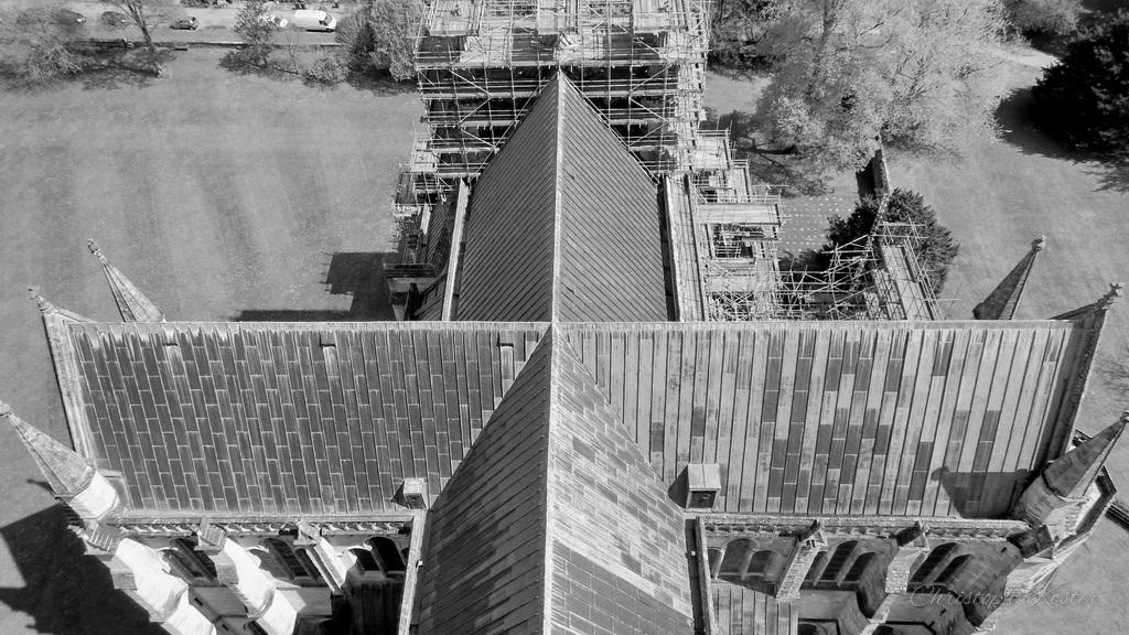 The image shows a black-and-white aerial view of a large church building, seen from directly above. The roof forms a cross-shaped layout, with long, sloping sections intersecting at the center.

The building features steep, textured roofing and symmetrical architectural elements, including rows of windows and decorative stone details along the sides. At the far end, part of the structure is covered in scaffolding, indicating ongoing construction or restoration work.

Surrounding the church are open grassy areas and a few trees, with a small number of parked cars visible in the background. The overall scene highlights the scale and structure of the historic building from a top-down perspective.