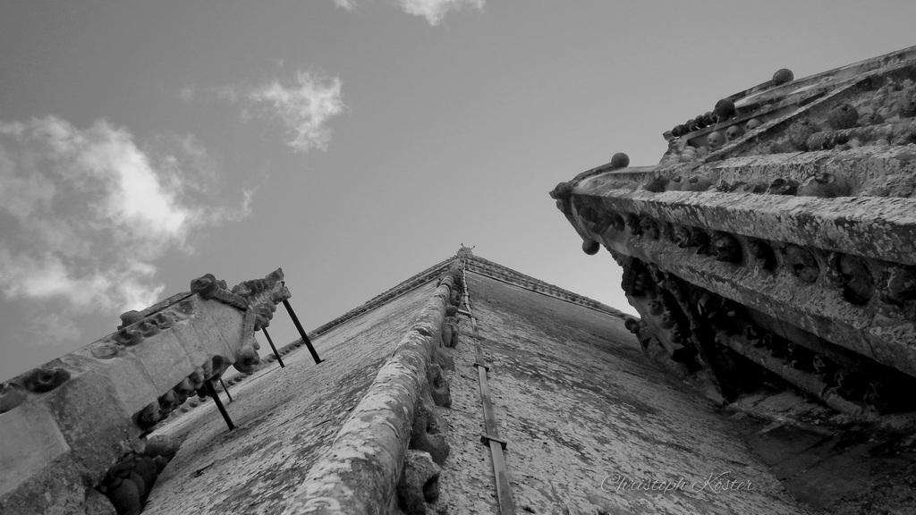 The image shows a dramatic black-and-white low-angle view of a historic stone building, likely a church or cathedral, looking straight up along its wall toward the roofline. The perspective emphasizes the building’s height and vertical structure, with the walls converging toward a narrow peak at the top.

On both sides, ornate stone details and decorative projections extend outward, including small sculptural elements and ledges. The surface of the wall appears weathered and textured, with visible aging and stone patterns.

Above, a partly cloudy sky forms the background, enhancing the sense of scale and drawing the eye upward along the architecture.