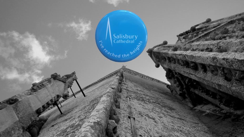The image shows a black-and-white low-angle view of a Gothic cathedral wall, looking sharply upward toward the roofline, which converges at a point high above. The stone surface is weathered and textured, with ornate architectural details and protruding decorative elements on both sides.

Above the building, a bright blue badge has been put artificially into the picture. The badge displays the text “Salisbury Cathedral – I’ve reached the heights” along with a simple graphic of a tall spire - it is the original badge you get, when you make the tower tour at Salisbury Cathedral.

The sky is lightly clouded, and the composition emphasizes the height of the structure.