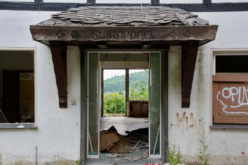 The image shows the entrance to an abandoned or derelict building with visible signs of decay. The focal point is a wooden canopy above the doorway, flanked by simple decorative carvings on both sides.

The building's facade is white stucco that appears weathered and stained, with visible cracks and peeling paint. The double wooden doors stand open, revealing a dark, empty interior space. Through the doorway, you can see a balcony or terrace with a metal railing, offering a view of green, rolling hills in the distance under a clear sky.

The tiled roof above the entrance is in poor condition, with some tiles missing or broken. On either side of the doorway are two windows with brown wooden frames:

    The left window has a broken or missing pane at the bottom.
    The right window is boarded up with a wooden shutter that has graffiti spray-painted on it, including the words 'OPERA' and 'VIVA'.

The ground in front of the entrance is uneven and cluttered, with debris including cardboard, plastic sheets, and other discarded materials. Weeds and small plants are growing along the base of the building, adding to the sense of abandonment.

The overall atmosphere of the image conveys neglect and disrepair, with the natural elements slowly reclaiming the space. The distant landscape provides a stark contrast to the dilapidated building, suggesting this might be a rural or semi-rural location.