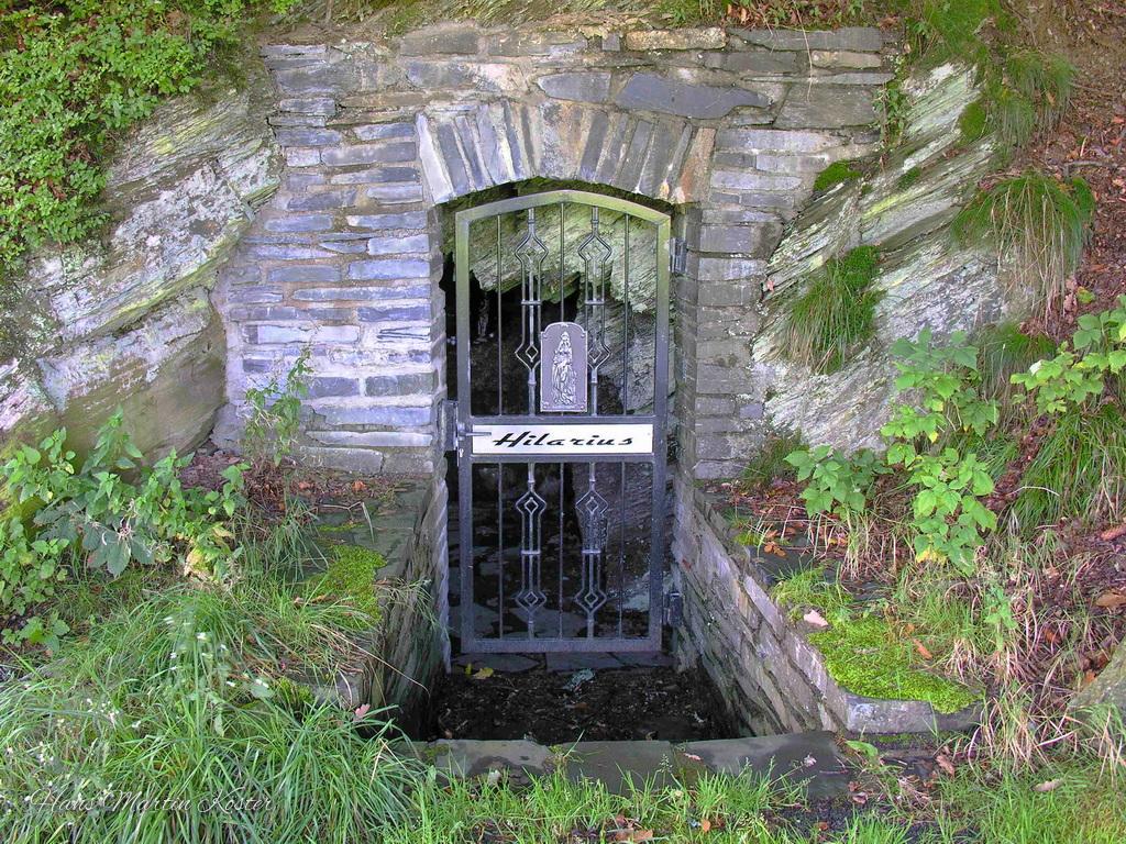The image shows a stone-built entrance set into a grassy hillside, framed by an arched structure made of stacked, weathered stones. A black metal gate with decorative geometric patterns blocks the doorway, which leads into a dark, narrow underground passage.

A white strip across the gate displays the word “Hilaritas.” The entrance is slightly recessed below ground level, with a shallow trench or step leading down to it.

Surrounding the doorway are green plants, grass, moss, and exposed rock, giving the scene a natural, overgrown look and a quiet, somewhat hidden atmosphere.
