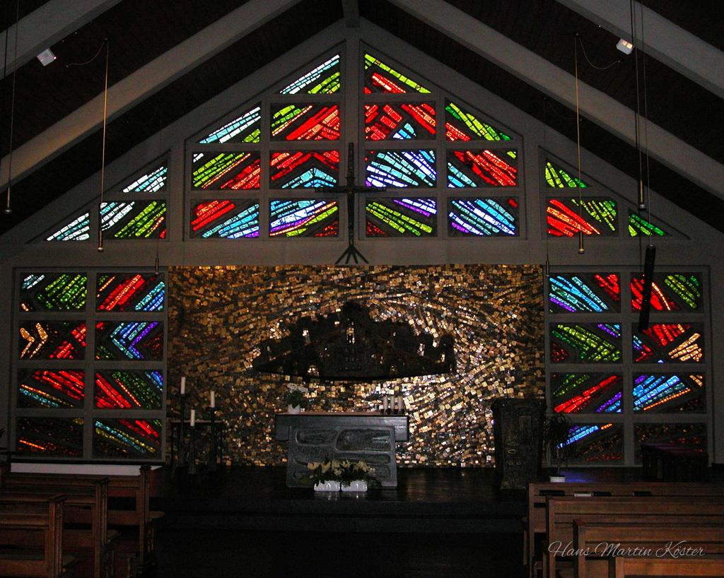 Das Bild zeigt den Innenraum einer Kirche mit modern gestalteten Buntglasfenstern. Im oberen Bereich sind dreieckig angeordnete Fensterflächen zu sehen, die in leuchtenden Farben wie Rot, Blau, Grün und Gelb schräg verlaufende Muster bilden.

Unterhalb der Fenster befindet sich eine golden schimmernde Mosaikwand, deren kleine, reflektierende Steine ein warmes Licht erzeugen. In der Mitte steht ein schlichter Altar aus Stein, darüber eine dunkle, abstrakte Form, die wie ein Baldachin oder eine Skulptur wirkt.

Im Vordergrund sind hölzerne Kirchenbänke zu erkennen, während die Decke durch sichtbare Balken und hängende Leuchten strukturiert ist. Die Szene vermittelt eine ruhige, feierliche Atmosphäre mit starkem Farbkontrast zwischen dem dunklen Raum und den hellen Glasfenstern.