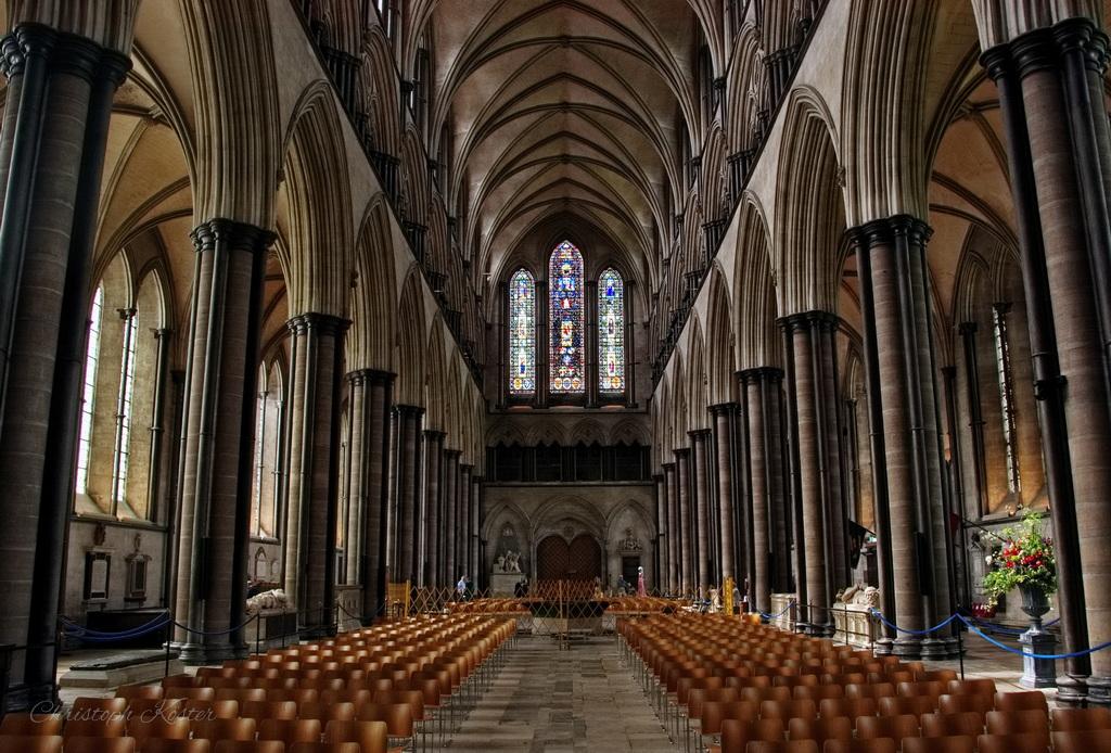 Interior of a large Gothic cathedral with high vaulted ceilings, rows of wooden chairs, tall stone columns, and stained glass windows at the far end.