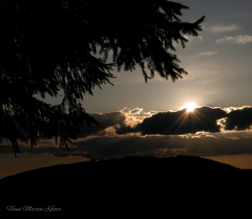 English:
Sun setting behind clouds over a dark hillside, with silhouetted tree branches in the foreground and rays of light breaking through the cloud cover.

Deutsch:
Sonnenuntergang hinter Wolken über einem dunklen Hügel, mit silhouettierten Baumzweigen im Vordergrund und Lichtstrahlen, die durch die Wolkendecke scheinen.

Français :
Coucher de soleil derrière des nuages au-dessus d’une colline sombre, avec des branches d’arbre en silhouette au premier plan et des rayons de lumière traversant la couverture nuageuse.